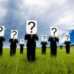 A group of people in black suits standing in a grassy field, each holding a large sign with a black question mark over their face.