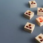 Wooden blocks with human icons are arranged on a gray surface; one block is highlighted in red, indicating emphasis or selection.