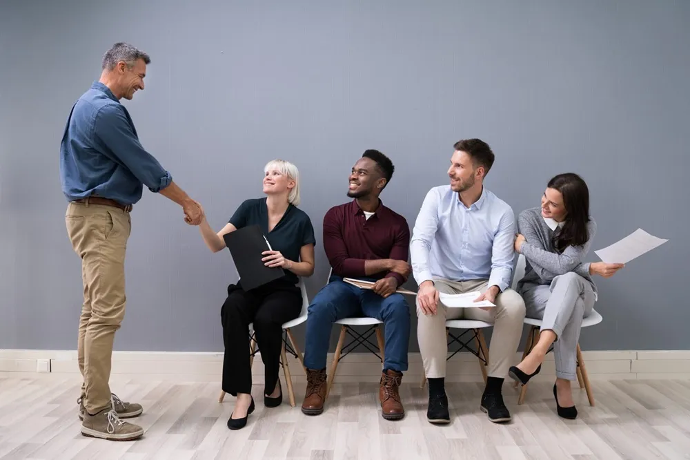 Five people in business casual attire are in an interview setting; one standing man shakes hands with a seated woman while three others watch and hold resumes.