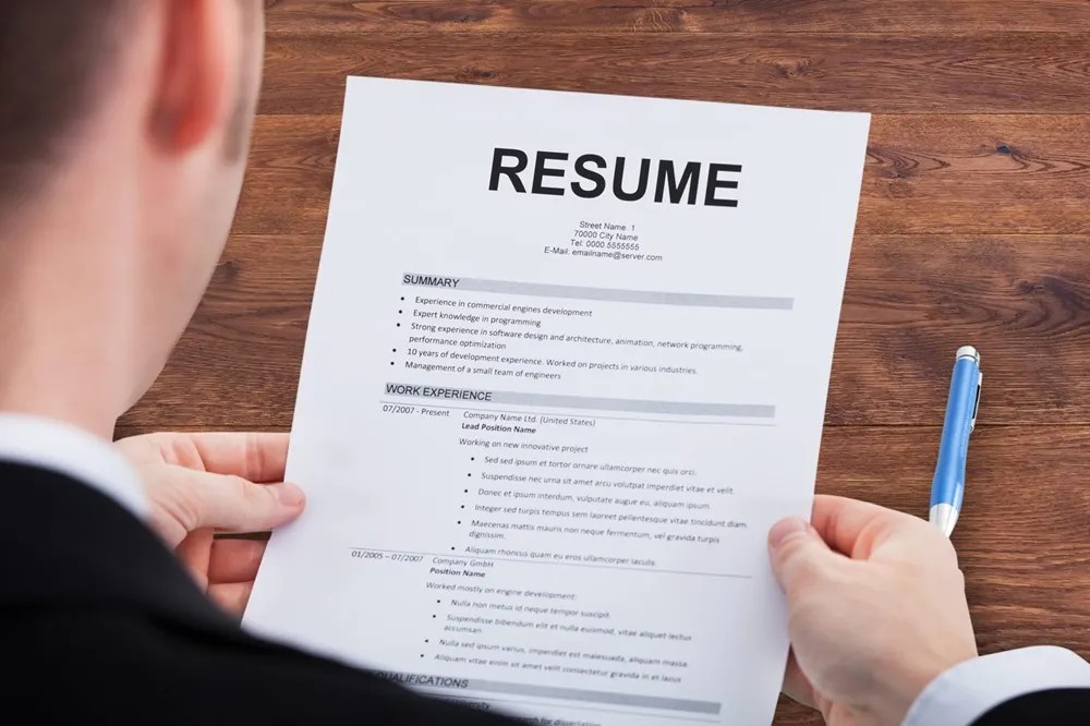 Person in a suit holding a printed resume document on a wooden desk, with a pen placed to the right of the paper.