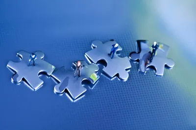 Four puzzle pieces are arranged in a line with miniature figurines of people standing on top of each piece against a blue background.