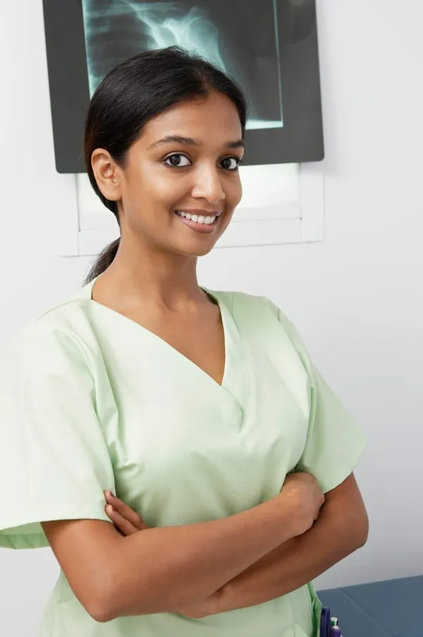 about A woman in light green scrubs stands with arms crossed, smiling, in a medical setting with an X-ray image visible in the background.